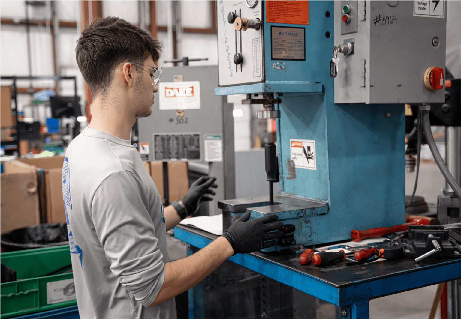 A man wearing safety glasses and gloves operates a blue industrial machine in Tekton's manufacturing center.