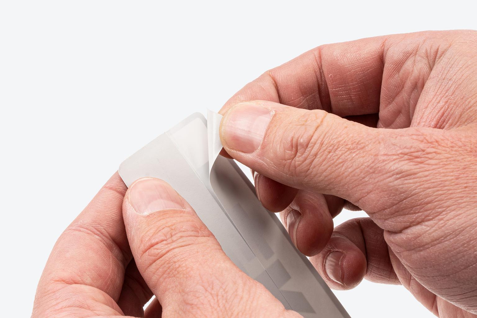 A hand pulling away half of the backer of a Tekton sticker, showing the scored backer.